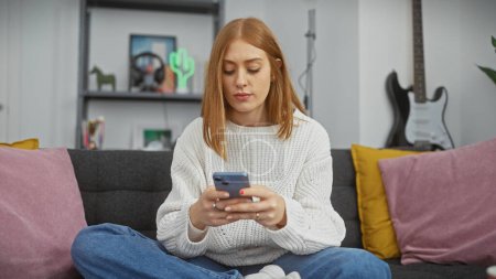 Mujer pelirroja serena mensajes de texto en el teléfono inteligente en un ambiente acogedor salón, ejemplificando el ocio interior moderno.