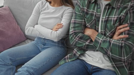 A man and woman sit with arms crossed on a sofa in a living room, portraying a couple possibly in a disagreement or contemplation.