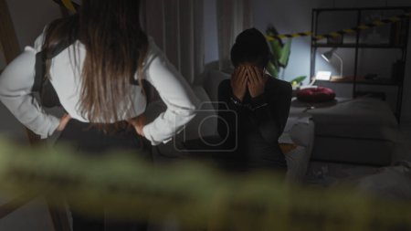 Téléchargez les photos : Une femme menottée assise avec découragement alors qu'une policière se tient derrière elle dans une pièce désordonnée et faiblement éclairée, bouclée avec une cassette de scène de crime. - en image libre de droit