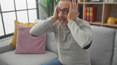Téléchargez les photos : Un homme d'âge moyen choqué avec une barbe dans des lunettes réagit émotionnellement alors qu'il est assis dans un salon moderne. - en image libre de droit