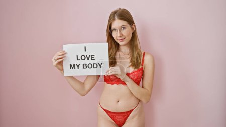 Foto de Joven confiada en lencería roja sosteniendo cartel con mensaje de positividad corporal contra telón de fondo rosa - Imagen libre de derechos