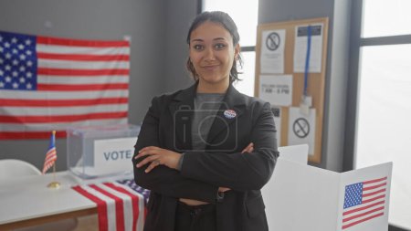 Mujer hispana joven con brazos cruzados se levanta orgullosamente en un centro de votación adornado con banderas americanas, retratando el compromiso cívico.