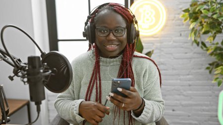 Photo for An african american woman with braids wearing headphones in a radio studio smiles while using a smartphone. - Royalty Free Image