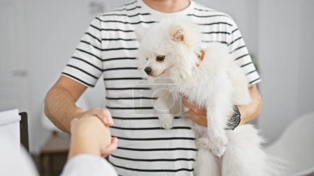 Handsome young caucasian man shaking hand with vet in a heartfelt greeting at the veterinary clinic's waiting room, lovingly looking at his puppy during consultation