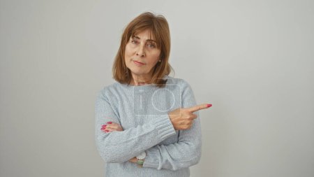 Téléchargez les photos : A sceptical middle-aged woman in a blue sweater points to the side against a white wall. - en image libre de droit