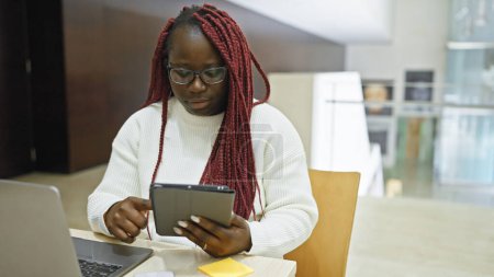 Téléchargez les photos : Femme afro-américaine focalisée avec des tresses en utilisant une tablette dans l'intérieur du bureau moderne. - en image libre de droit