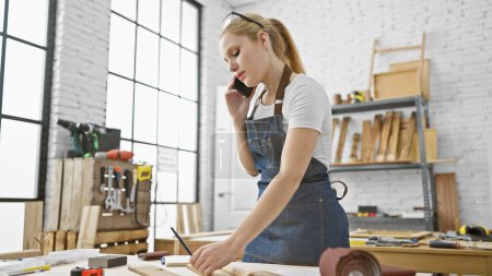 Foto de Una mujer rubia en un delantal de mezclilla habla por teléfono en un brillante taller de carpintería lleno de herramientas y tablones de madera. - Imagen libre de derechos
