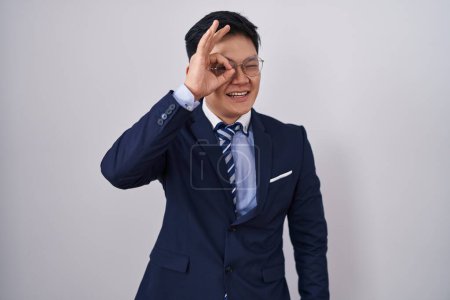 Foto de Joven asiático hombre vistiendo traje de negocios y corbata haciendo ok gesto con la mano sonriente, ojo mirando a través de los dedos con cara feliz. - Imagen libre de derechos
