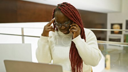 Photo for A concentrated african american woman with braids talking on the phone in a modern office setting. - Royalty Free Image
