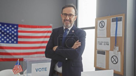 Foto de Hombre maduro de confianza en traje con los brazos cruzados de pie en un centro de votación con bandera americana - Imagen libre de derechos