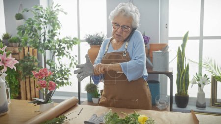 Foto de Una mujer mayor con guantes prepara flores en una brillante tienda de jardín interior, que encarna la madurez y el cuidado. - Imagen libre de derechos