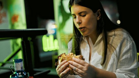 Téléchargez les photos : Jeune belle femme hispanique mangeant un délicieux hamburger au restaurant - en image libre de droit