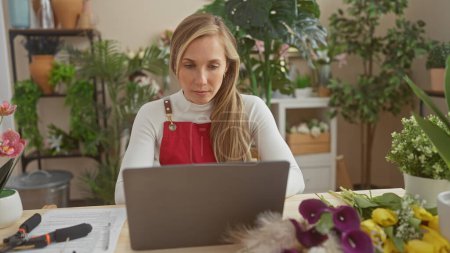 Téléchargez les photos : Une femme caucasienne concentrée travaillant sur un ordinateur portable entouré de fleurs vibrantes et de verdure luxuriante dans un cadre de fleuriste d'intérieur. - en image libre de droit