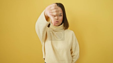 Photo for Hispanic young woman with neutral expression wearing sweater against yellow wall giving thumbs down gesture. - Royalty Free Image