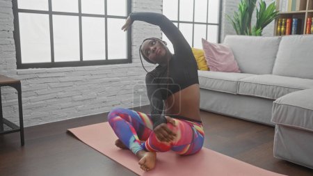 Foto de Mujer afroamericana estirándose sobre una esterilla de yoga en una moderna sala de estar con un sofá en el fondo. - Imagen libre de derechos