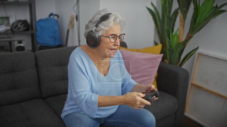 Téléchargez les photos : Femme âgée avec écouteurs jouant à des jeux vidéo dans un salon confortable, mettant en valeur le mode de vie actif des personnes âgées - en image libre de droit