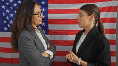 Foto de Dos mujeres profesionales conversando sobre un telón de fondo de una bandera americana en el interior de una oficina. - Imagen libre de derechos
