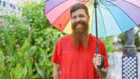 Foto de Joven pelirrojo alegre disfrutando con confianza de un día lluvioso en el parque de la ciudad, paraguas en la mano, sonrisa guapo irradiando alegría y vibraciones positivas - Imagen libre de derechos