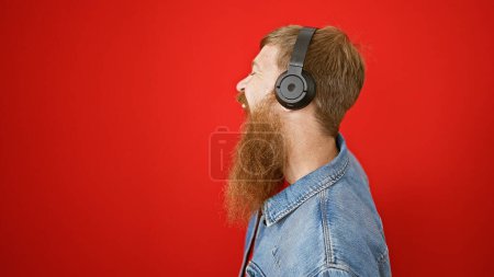 Foto de Joven pelirrojo escuchando música haciendo un gesto hacia arriba sobre un fondo rojo aislado - Imagen libre de derechos