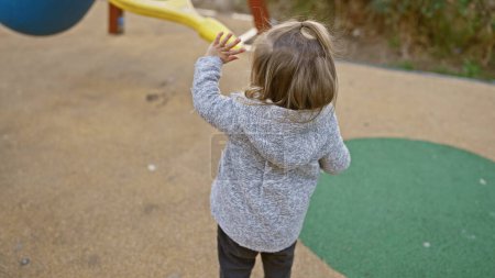 Photo for Blonde girl playing outdoors on a colorful playground, reaching towards a slide in a park - Royalty Free Image