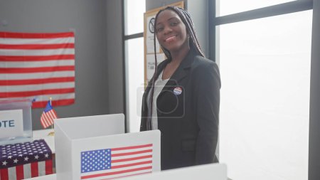 Photo for Portrait of a confident african american woman with braids wearing a 'voted' sticker indoors at a polling station with american flags. - Royalty Free Image