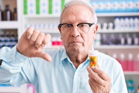 Téléchargez les photos : Homme âgé avec les cheveux gris tenant des pilules à la pharmacie avec le visage en colère, signe négatif montrant aversion avec les pouces vers le bas, concept de rejet - en image libre de droit