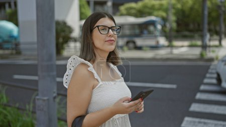 Foto de En las bulliciosas calles de Tokyo, una hermosa chica hispana con gafas, una viajera conocedora de la tecnología, utiliza su aplicación de teléfono, esperando ansiosamente su viaje en taxi. - Imagen libre de derechos