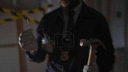 Téléchargez les photos : Un homme afro-américain inspecte les preuves sur une scène de crime à l'intérieur, tenant un marteau et portant des gants. - en image libre de droit