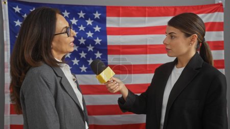 A woman reporter interviews another woman against a background of the american flag indoors.