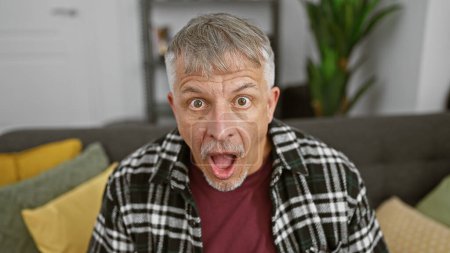 Foto de Hombre maduro sorprendido con el pelo gris en el interior de una casa, expresando conmoción y emoción. - Imagen libre de derechos