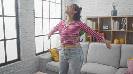 Foto de Joven mujer hispana alegre bailando en una acogedora sala de estar con decoración moderna. - Imagen libre de derechos