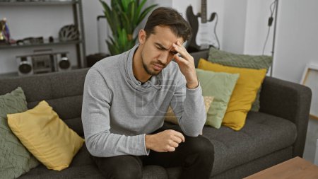 Foto de A young hispanic man with a beard appears stressed while sitting on a gray couch indoors, showcasing a modern home interior. - Imagen libre de derechos