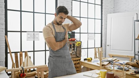 Foto de Hombre hispano guapo con barba en el taller usando delantal sobre ropa casual - Imagen libre de derechos
