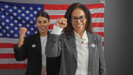 Téléchargez les photos : Deux femmes aux poings levés en victoire ou solidaires devant un drapeau américain, à l'intérieur, portant des autocollants "j'ai voté" - en image libre de droit