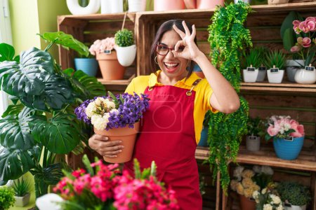 Foto de Mujer árabe joven que trabaja en floristería sosteniendo la planta sonriendo feliz haciendo signo bien con la mano en el ojo mirando a través de los dedos - Imagen libre de derechos
