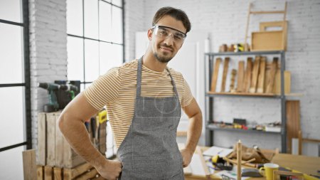 Foto de Un joven hispano confiado con barba con gafas de seguridad y un delantal posa en un taller de carpintería bien equipado. - Imagen libre de derechos