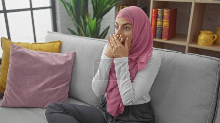 Foto de Mujer joven sorprendida usando un hijab sentado en un sofá interior con almohadas de colores y fondo de estantería - Imagen libre de derechos