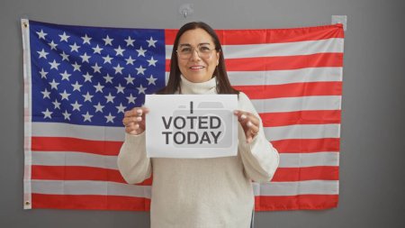 Foto de Mujer hispana de mediana edad sostiene el letrero 'Hoy he votado' ante la bandera americana, retratando el deber cívico en un ambiente interior. - Imagen libre de derechos