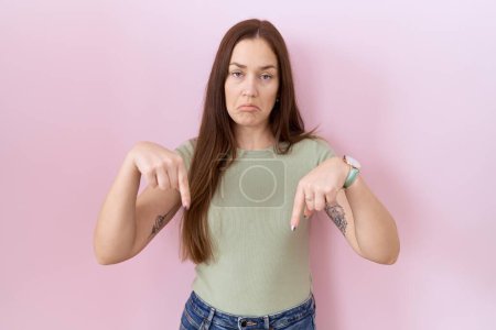 Schöne brünette Frau, die vor rosa Hintergrund steht und traurig und aufgebracht nach unten zeigt, die Richtung mit den Fingern anzeigt, unglücklich und deprimiert. 