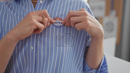 Caucasian woman in a striped shirt pins a 'voted today' button, symbolizing american democratic participation indoors.