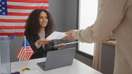 A woman officiates at a usa electoral event, handing papers to a man in front of an american flag inside a room.