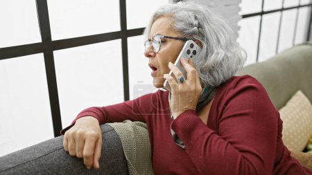 A mature woman with grey hair talks on the phone while sitting on a sofa indoors, showing concern and urgency.