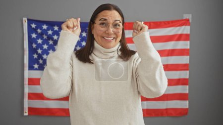 Eine fröhliche hispanische Frau mittleren Alters feiert in einem Büro mit US-Flaggenhintergrund.