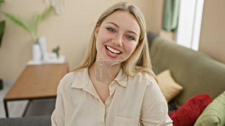 Foto de Mujer rubia sonriente en una camisa beige casual posando cómodamente en su moderna sala de estar amueblada. - Imagen libre de derechos