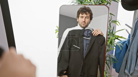 Téléchargez les photos : Un beau jeune homme avec une barbe essayant un costume dans un dressing, reflété dans un miroir - en image libre de droit