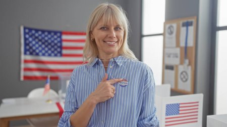 A smiling caucasian woman in a striped shirt shows her 'i voted' sticker with american flags in a college polling center.