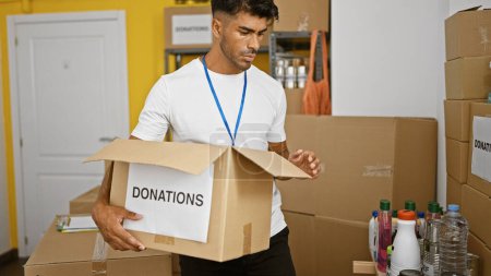 Téléchargez les photos : Un bel homme hispanique faisant du bénévolat dans un entrepôt, portant une boîte de dons au milieu de fournitures. - en image libre de droit