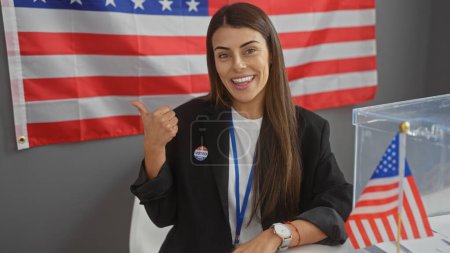 Foto de A young hispanic woman with an 'i voted' sticker, pointing to the american flag at an electoral college. - Imagen libre de derechos