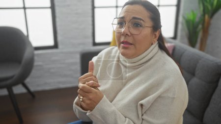 Photo for A mature hispanic woman with glasses sits thoughtfully in a cozy living room, expressing a point during a conversation. - Royalty Free Image