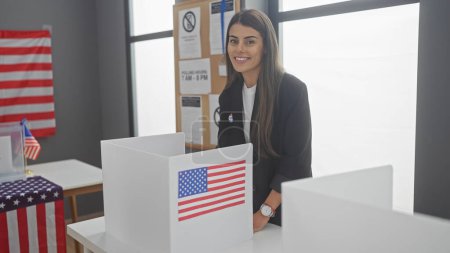 Una joven hispana sonríe en un centro electoral americano con banderas e información electoral.
